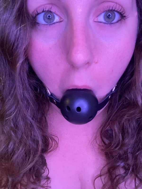 Close-up portrait of a woman with curly brown hair and blue eyes, wearing a black leather accessory around her mouth against a dark background.