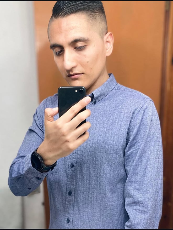 Young man with short dark hair wearing a light blue button-up shirt, holding a smartphone, standing indoors against a wooden door.