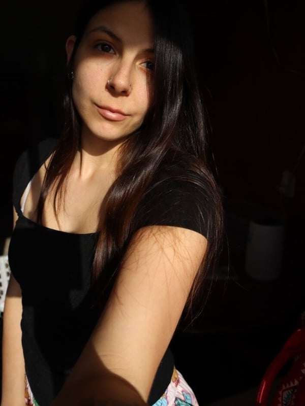 Young woman with long dark hair smiling softly at the camera, wearing a black off-shoulder top and colorful pants in a dimly lit room.