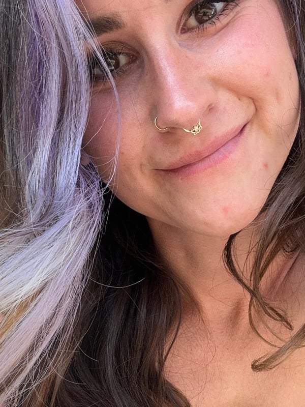 A smiling woman with purple-streaked hair and a nose piercing, standing near a door with tattoos visible on her shoulder.
