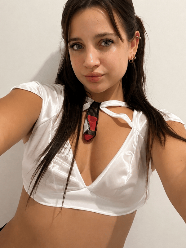 A young woman taking a selfie in a white satin crop top with a small tie detail, against a plain wall.