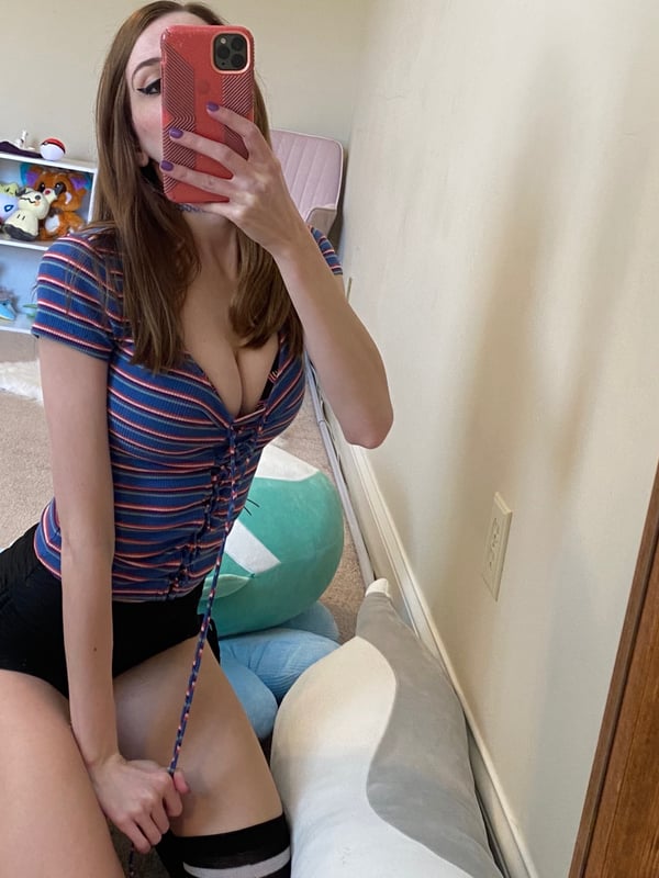 A woman taking a mirror selfie in a striped top and skirt, holding a phone, with toys and decorations in a cozy room background.