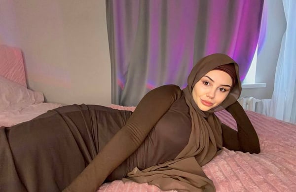 A woman in modest brown attire and headscarf reclining on a pink bed in a bedroom with purple lighting and curtains.