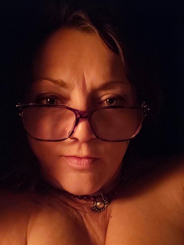 Close-up portrait of a woman with glasses and a necklace, looking directly at the camera against a dark background.