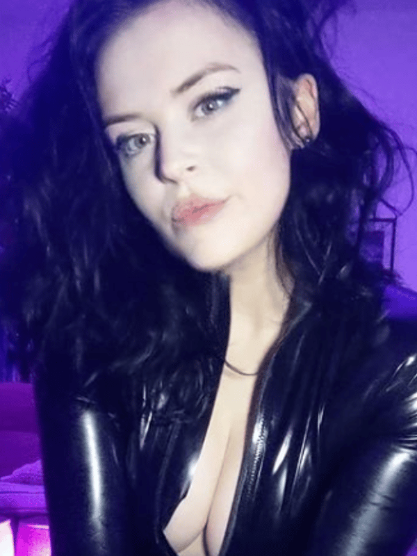 Close-up portrait of a woman with wavy black hair and bold makeup, wearing shiny black attire in purple lighting.