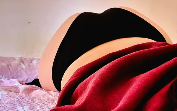 A person lying on a bed with pink sheets, wearing black underwear and a red blanket draped over their lower body.