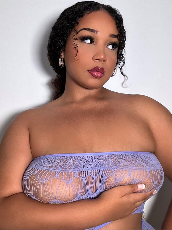 Close-up portrait of a woman with curly black hair, wearing sheer purple lingerie, posing with hands on her chest against a white background.