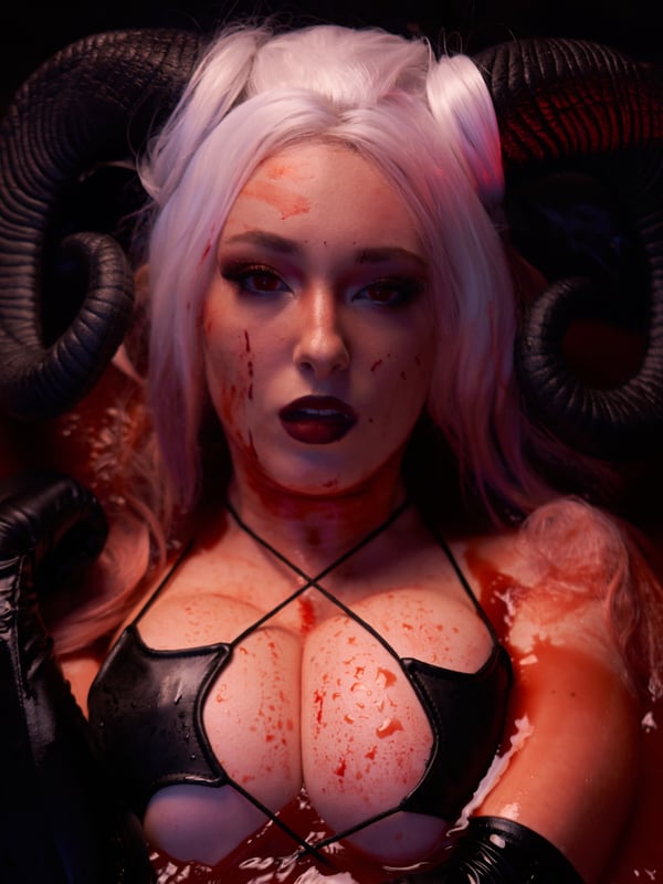 A person in a horned costume with white hair, wearing black leather straps, posing dramatically in a red liquid setting with rose petals.