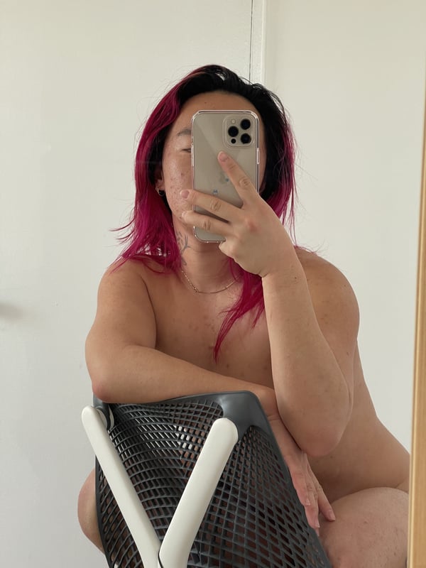 Person with pink hair taking a mirror selfie seated on a chair, appearing shirtless in a casual home setting.