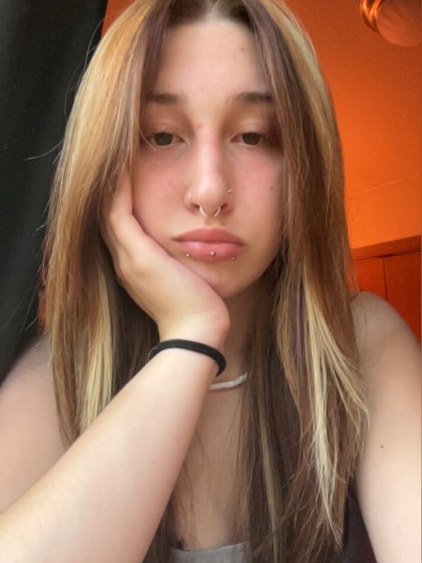 Close-up of a young woman with long highlighted hair, resting her hand on her cheek, wearing a gray top against an orange-lit background.