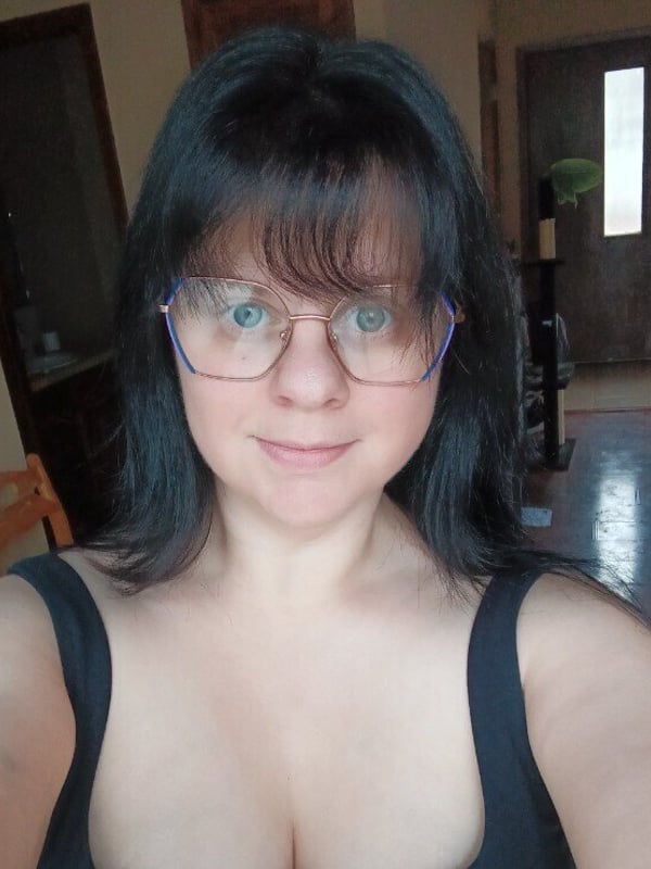 Woman with dark hair and glasses smiling in a black top inside a home with wooden doors and furniture.