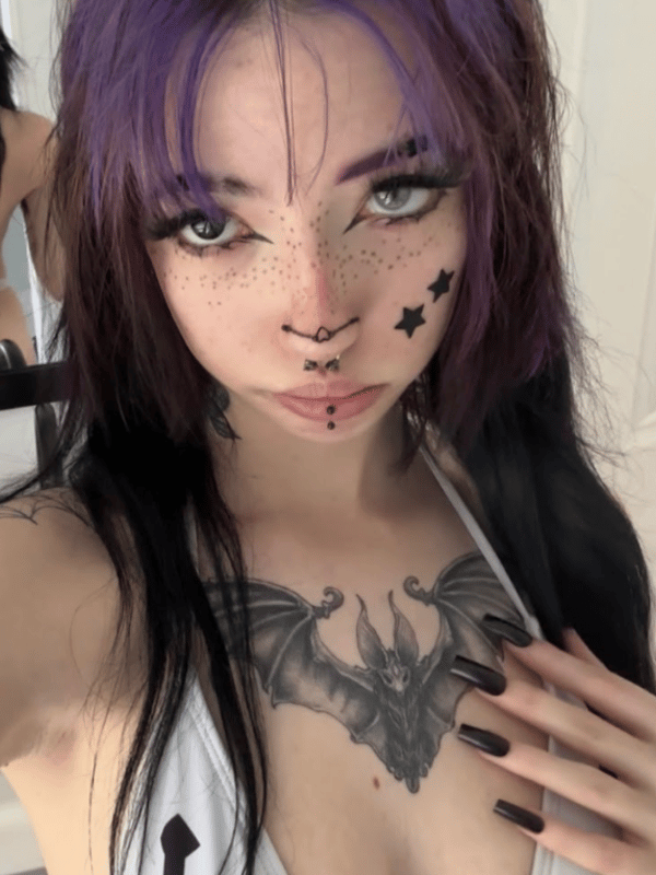A close-up selfie of a person with purple and black hair, facial piercings, and a large bat tattoo on the chest.
