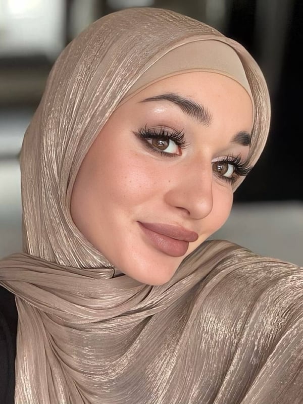 Close-up selfie of a woman with a warm smile, wearing a shiny beige hijab and subtle makeup, against a softly lit indoor background.