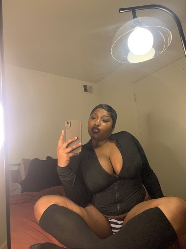A woman taking a mirror selfie in a bedroom, wearing a black zip-up top and striped stockings while sitting on a bed.