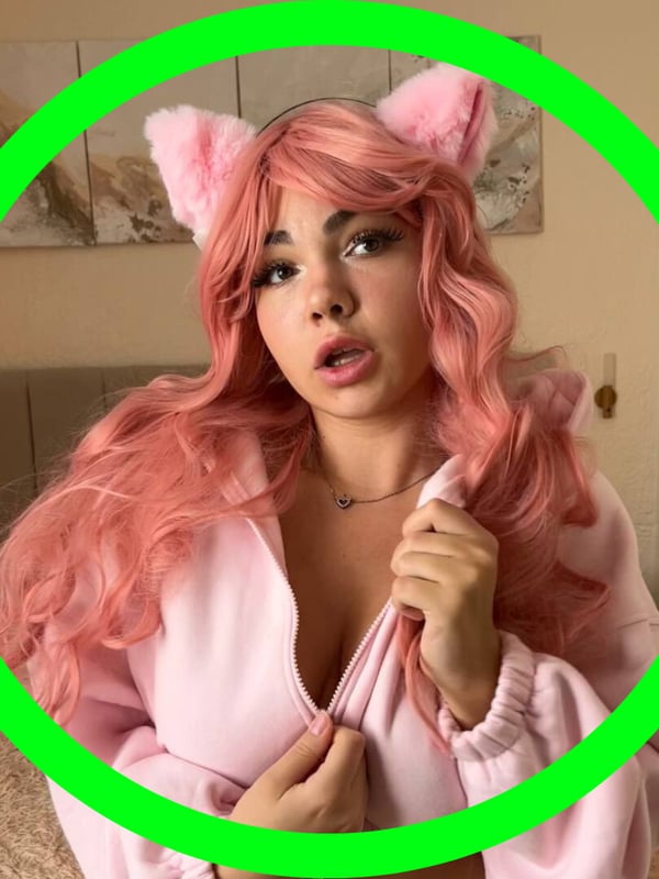 Close-up of a young woman with pink hair, cat ears, and a surprised expression wearing a pink hoodie.