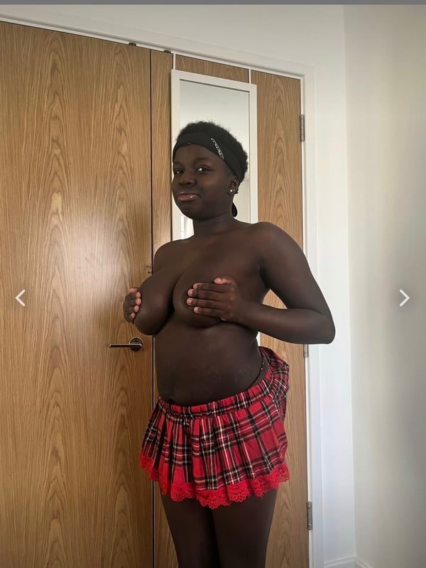 Young woman with dark skin standing in front of wooden doors, covering her chest with her hands, wearing a short red plaid skirt.