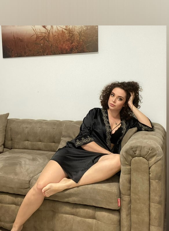 A woman with curly hair lounging on a couch in a silky black robe, with a scenic painting in the background.