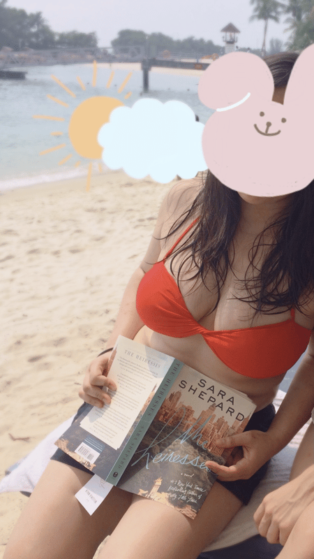 A young woman in a red bikini sits on a beach chair holding a book, with a scenic waterfront and palm trees in the background, overlaid with a cute bunny sticker on her face.