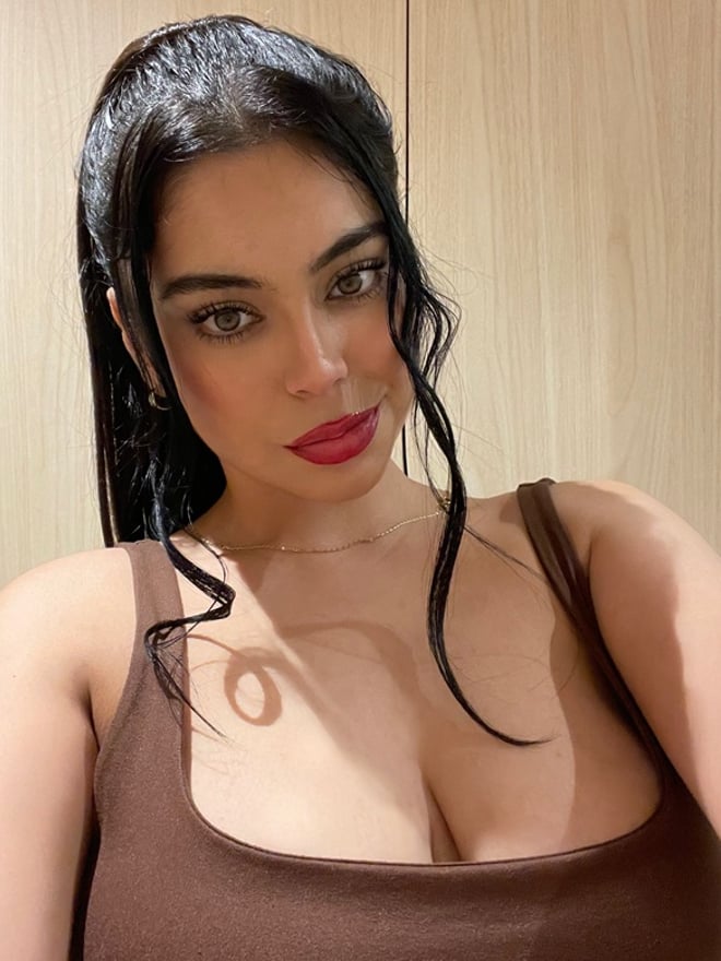 A woman with long dark hair wearing a red lace bra, posing against a plain background while looking at the camera.