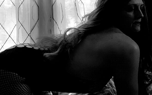 A woman with long hair poses sideways near a window with sheer curtains in black and white.