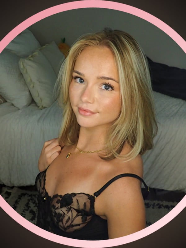 Close-up of a smiling young woman with blonde hair and blue eyes, wearing black lace top, framed in a pink circle against a cozy bedroom background.