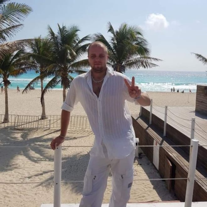 Man with bald head and goatee wearing white shirt and pants, posing with peace sign on a beach deck surrounded by palm trees and ocean.