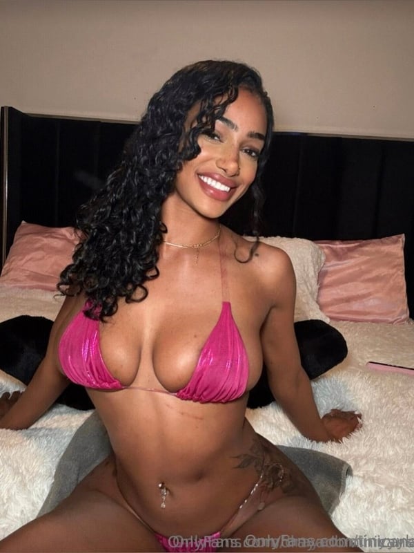 Young woman with curly black hair and freckles smiling on a bed, wearing a pink bikini top and jewelry.