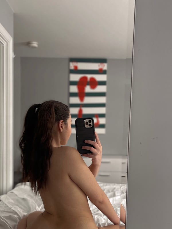 A woman taking a mirror selfie in a bedroom, holding her phone while seated on a bed.