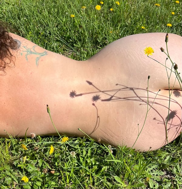 A woman lying on her side in a grassy field surrounded by yellow flowers, with a tattoo visible on her back.