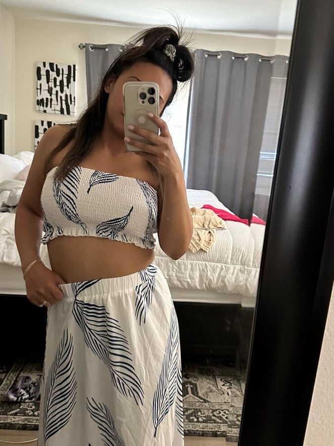 A woman taking a mirror selfie in a bedroom, wearing a white floral tube top and skirt.