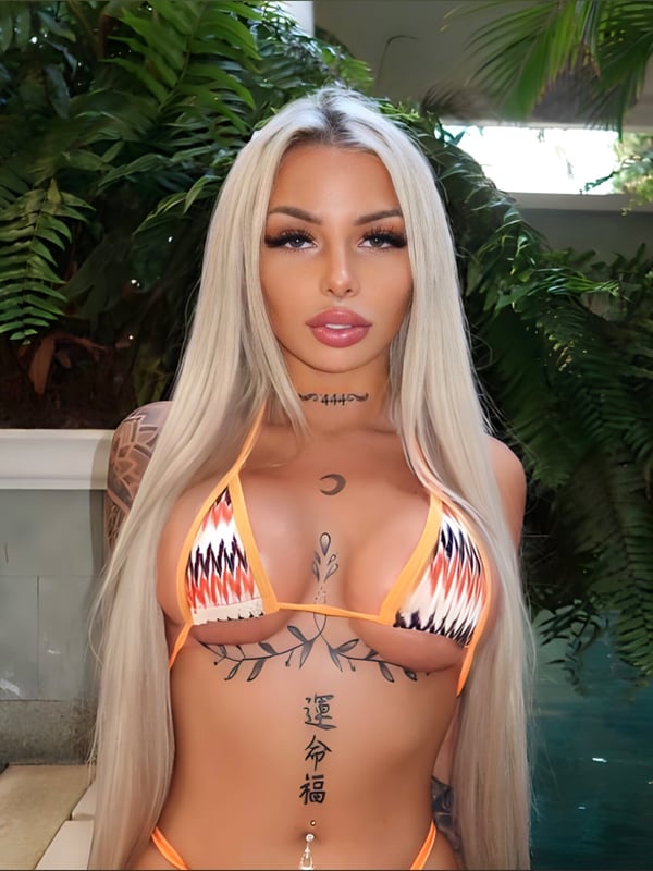 A woman with long blonde hair wearing an orange bikini top, standing near tropical plants and water.