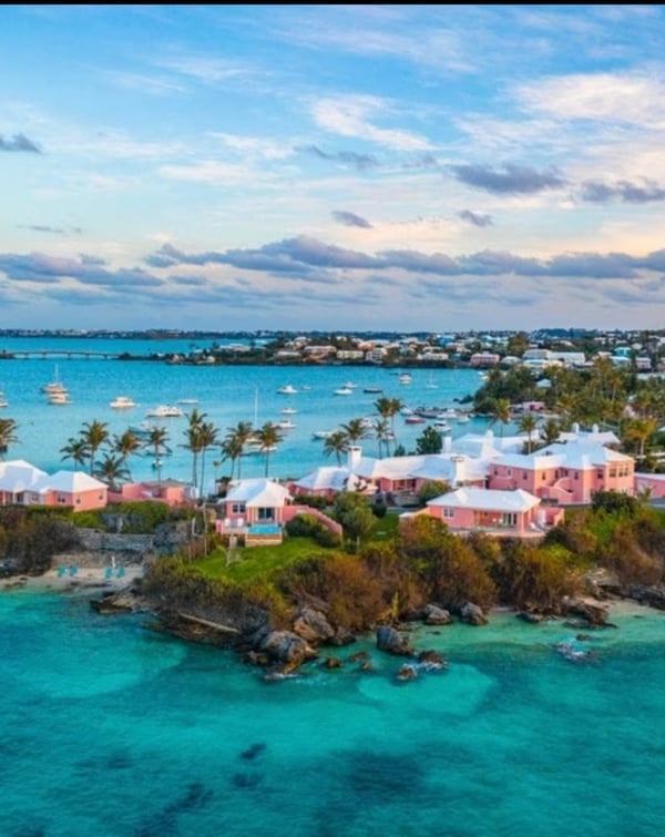 Aerial view of a scenic coastal area with pink buildings, turquoise waters, yachts, and palm trees at sunset.