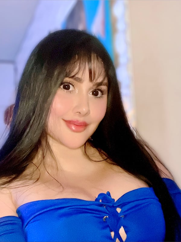 Young woman with long dark hair and bangs smiling at the camera, wearing a blue off-shoulder top, standing indoors near a window.