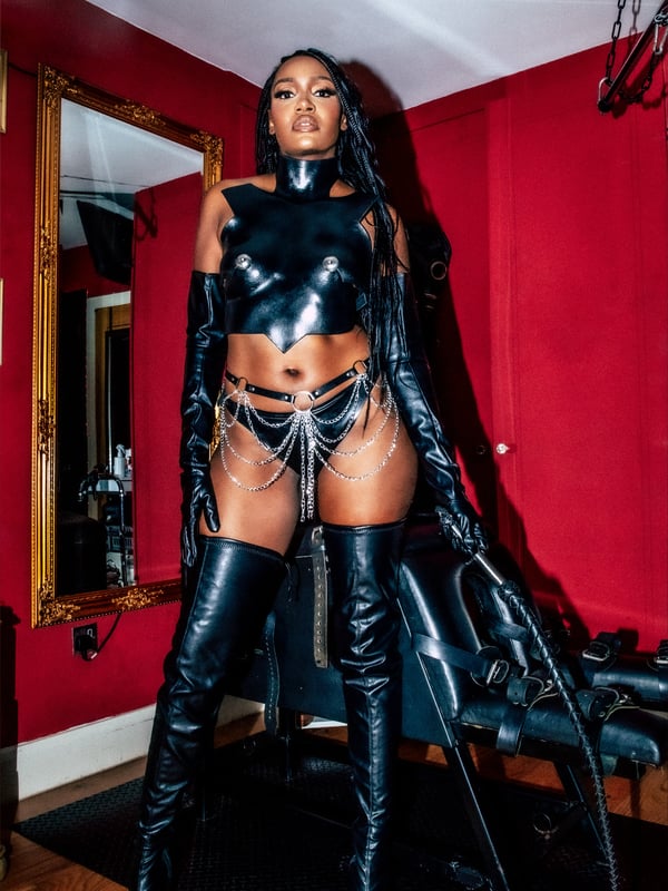 A woman in a dominant pose wearing latex outfit, long boots, and holding a whip in a dimly lit room with BDSM decor.