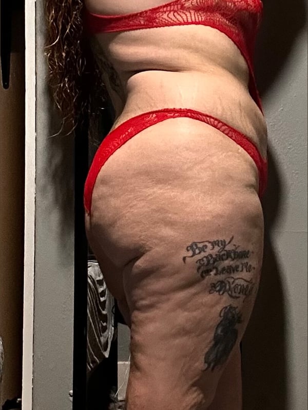 A woman with curly hair posing sideways in red lingerie in front of a mirror, showing tattoos on her arms and legs.