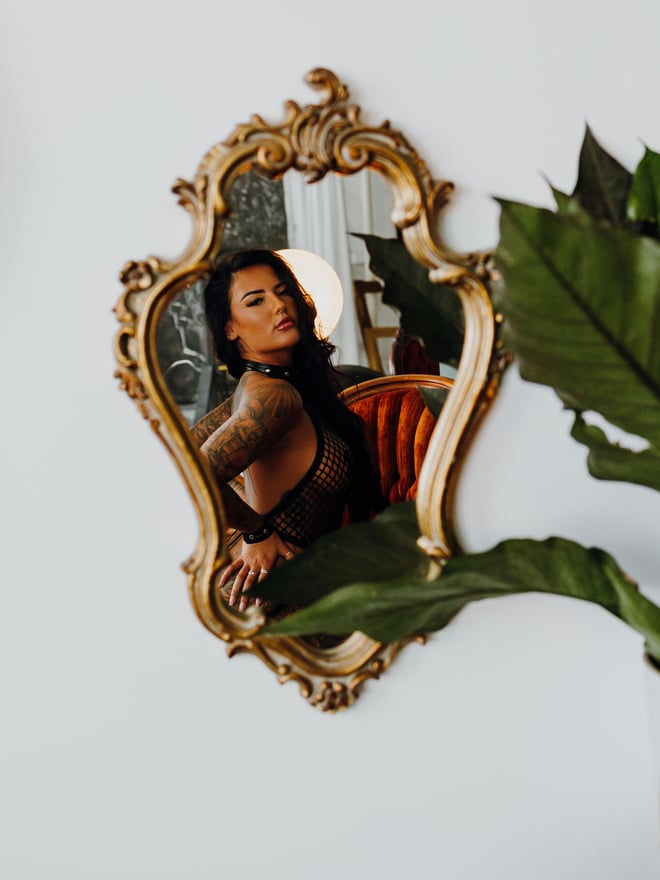 Close-up reflection of a tattooed woman in an ornate gold mirror frame with plants and a chair nearby.