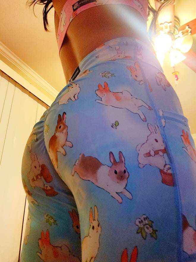 A person standing in a room with warm lighting, wearing colorful bunny-patterned leggings that highlight their figure.