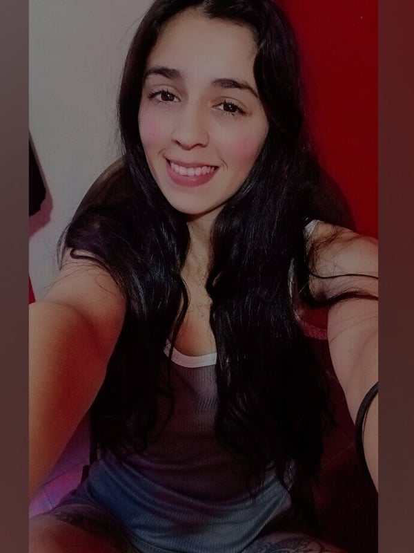 A young woman with long dark hair smiling at the camera in a close-up selfie, wearing a gray tank top, against a reddish background.