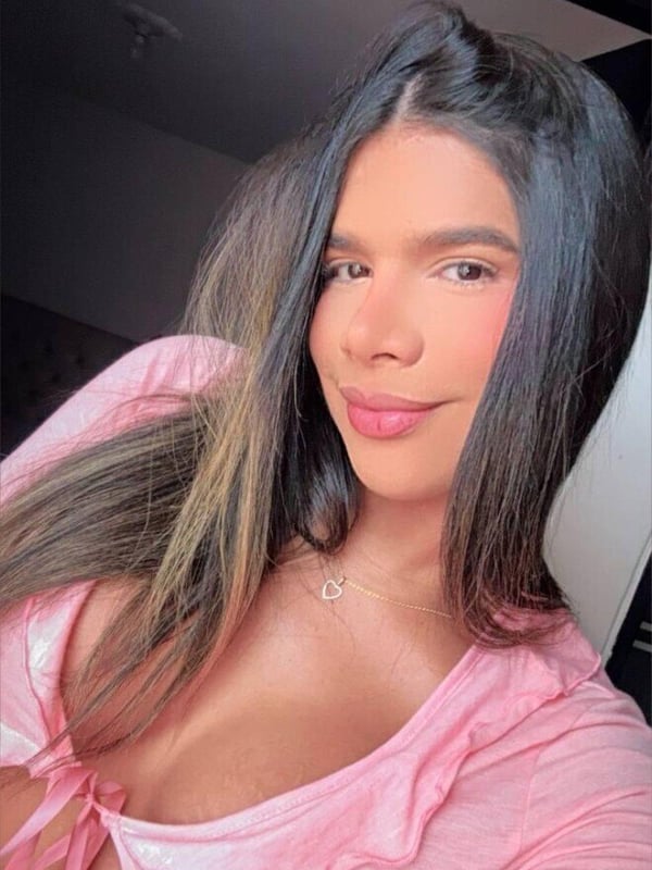 A close-up portrait of a person with long dark hair, wearing a pink top, smiling softly indoors.