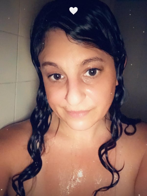 A woman with wet curly black hair smiling in a bathroom with tiled walls.
