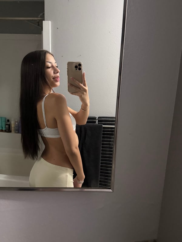 Young woman with long dark hair posing in a zebra-print bodysuit and fluffy boots, taking a mirror selfie in a bedroom with warm lighting.