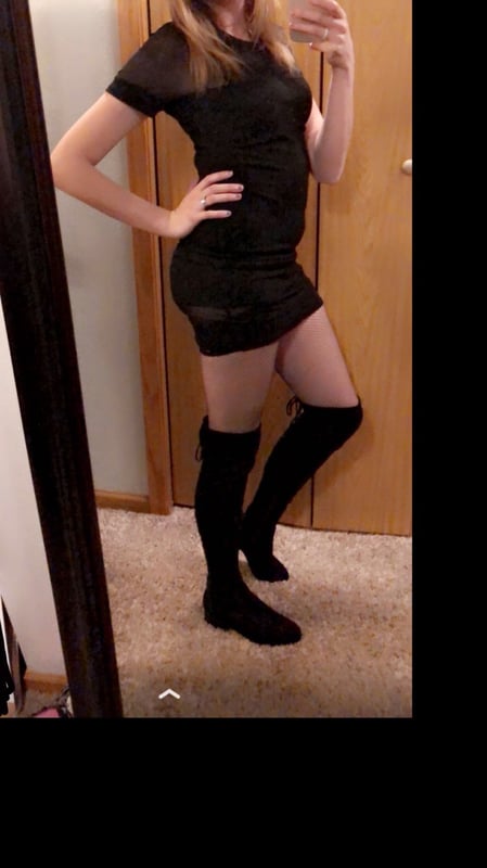 Young woman with long blonde hair posing in a short black sheer dress and over-the-knee boots in front of a mirror inside a room with wooden doors.
