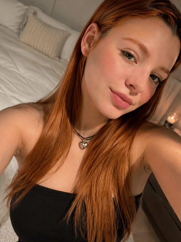 A young woman with long reddish-brown hair taking a selfie in a bedroom while wearing a black top and necklace.