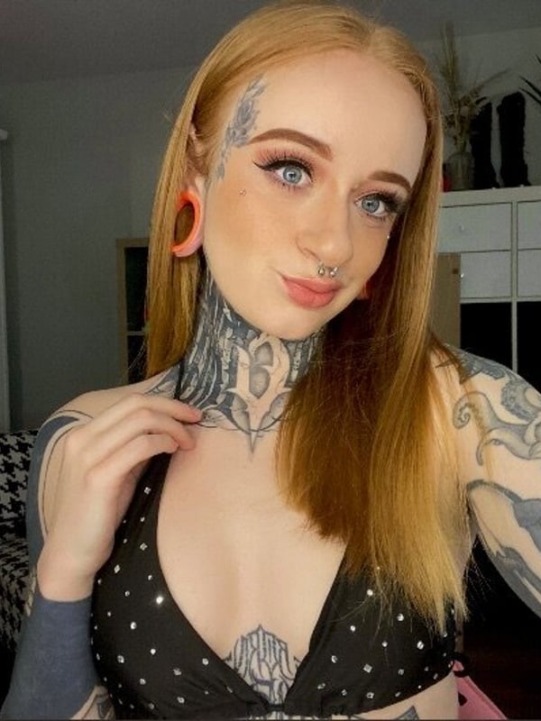 Young woman with long blonde hair, tattoos, and large earrings smiling at the camera in a bedroom setting with plants and lighting equipment.