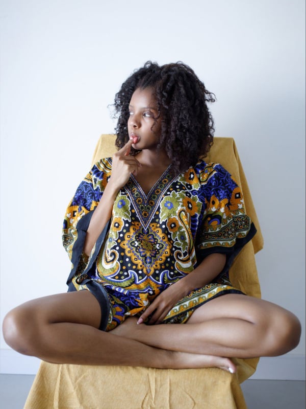 A woman with curly hair in a colorful patterned dress sits on a yellow chair with legs crossed, posing thoughtfully against a simple background.