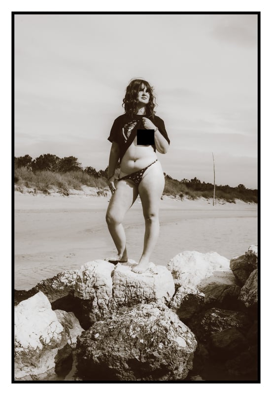 A woman stands confidently on rocks by the beach in a black and white photo, wearing a t-shirt and bikini bottoms with a scenic ocean backdrop.