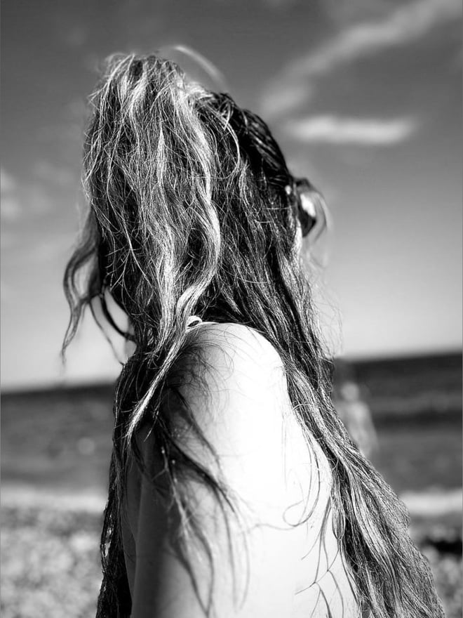 Black and white profile silhouette of a person with long wavy hair standing on a rocky beach with ocean waves and cloudy sky in the background.