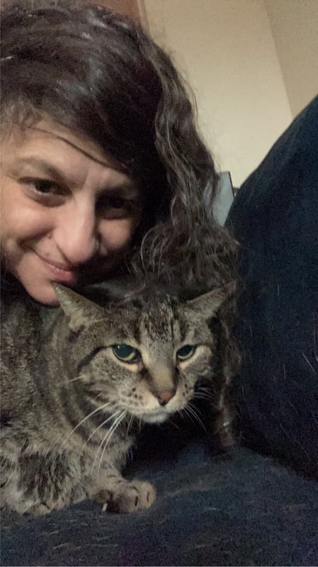 A woman with curly brown hair smiling while cuddling a tabby cat on a dark couch.