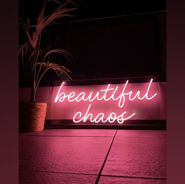 Neon sign reading 'beautiful chaos' in pink lighting next to a potted plant on a tiled floor with pink hues.