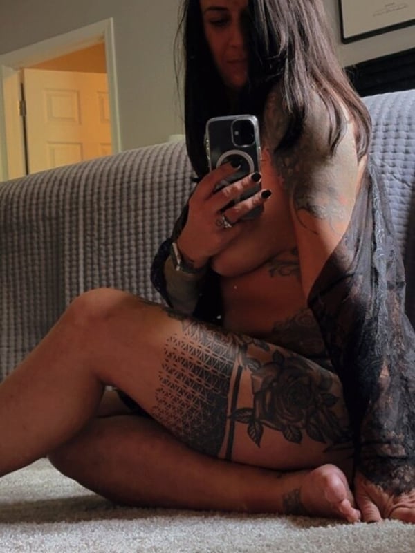 A woman taking a selfie in a cozy indoor setting with a record on the wall, wearing a sheer top.